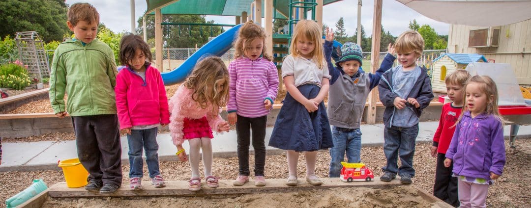 A group of young children standing in a line at the edge of a sandbox in an outdoor playground, with various playground equipment and greenery in the background. The children are dressed in colorful jackets and appear to be engaged in an activity.