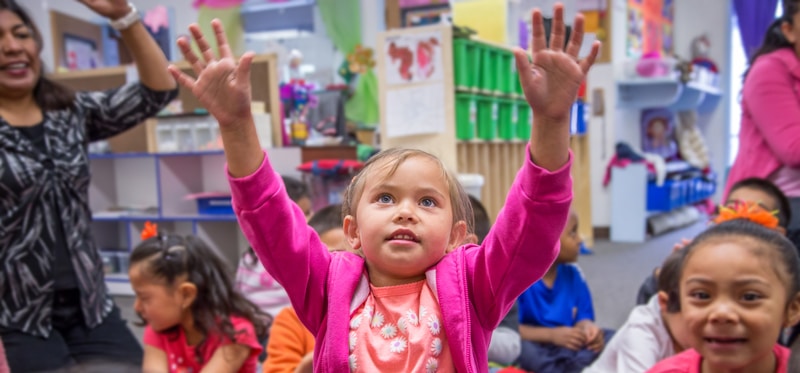 A young girl in a pink jacket raises her hands enthusiastically while sitting with other children in a colorful classroom. A teacher in the background also raises her hands, engaging the children in an interactive activity.