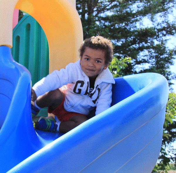 A young child with curly hair, wearing a white jacket and red shorts, smiles while playing on a colorful playground slide on a sunny day, with trees in the background.