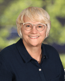 An older person with short, light blond hair and glasses smiles at the camera. They are wearing a dark blue shirt and a necklace. The background is blurred with green foliage, suggesting an outdoor setting.