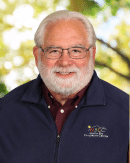 A smiling older man with white hair and a beard wearing glasses, a maroon shirt, and a navy blue jacket. The background is outdoor with blurred green and yellow foliage.